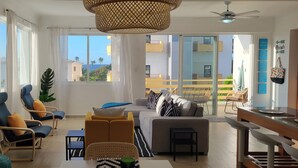 Uma sala de estar moderna com sofá, poltronas, uma mesa de jantar e uma varanda com vista para edifícios e palmeiras.