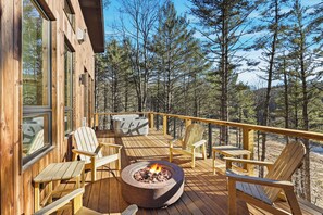 Um deck de madeira com cadeiras Adirondack, uma lareira e vista para uma floresta.