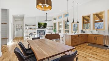 A modern kitchen with wooden cabinets, a dining area with a large table and chairs, and a view of the outdoors through large windows.