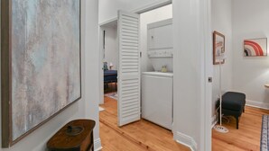 A hallway with a wooden floor, a white door leading to a room with a bed, a washing machine, and a small table with a lamp. - 2Blocks to Jazzfest Gate! Spotless 3BR w Driveway (New Orleans)