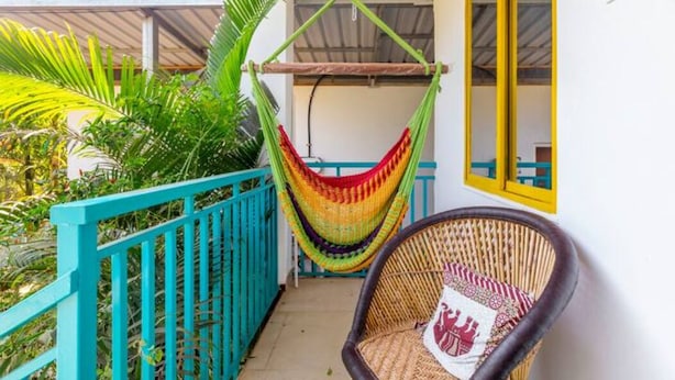 A balcony with a hammock, a wicker chair, and a view of a building with a yellow window frame. - Zennis Earth Stays (Mandrem)