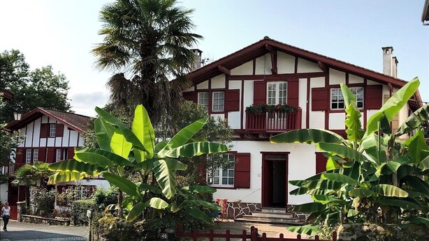 Een huis met twee verdiepingen en een balkon, omringd door weelderige begroeiing en een stenen muur.