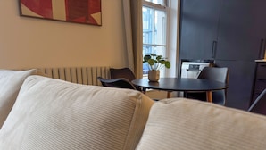 A modern living room with a sofa, a small table with a potted plant, and a view of a kitchen and outside through a window. - Large Luxury Apartment Combo featuring 4 bedrooms, 4 bathrooms & 2 living rooms. (London)
