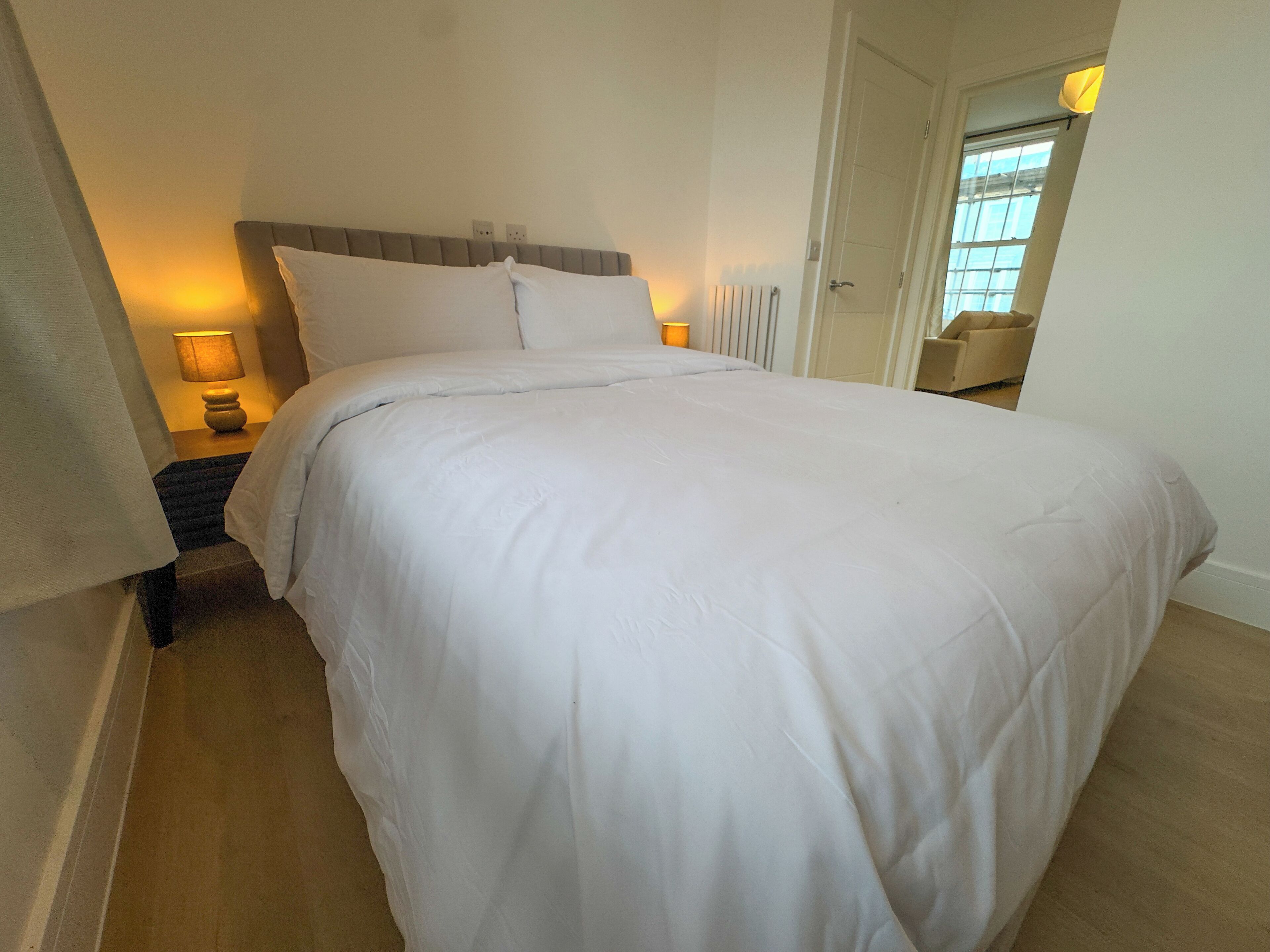 Een net opgemaakt bed in een hotelkamer, met een hoofdbord, bedlampjes en een raam met zonwering.