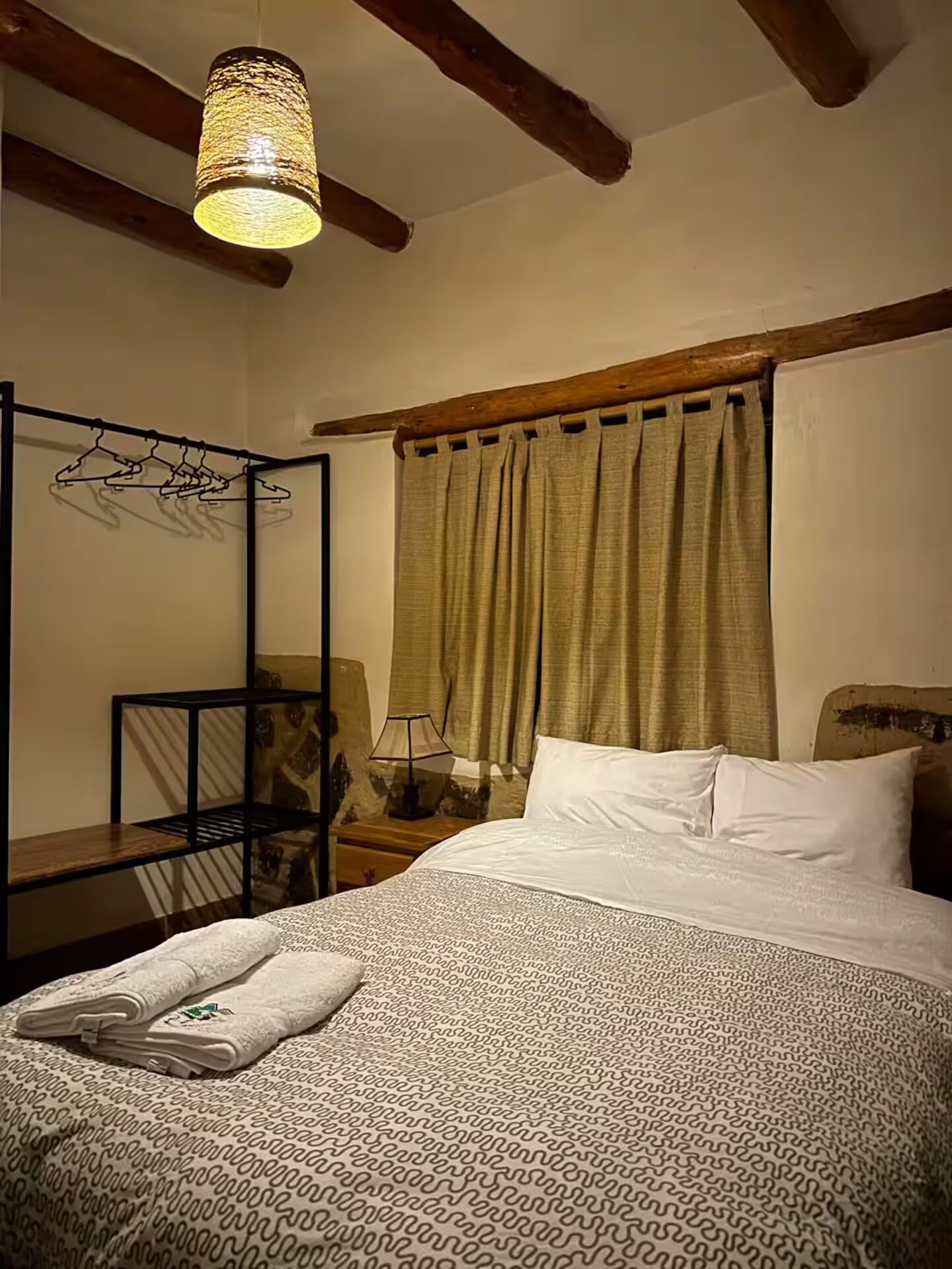 A bedroom with a bed, a hanging light fixture, a wooden headboard, a nightstand, a chair, and a window with curtains.