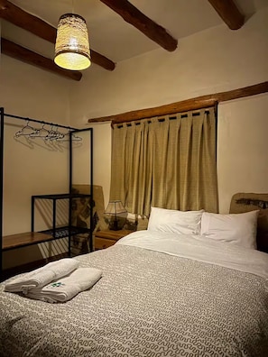 Ein Schlafzimmer mit Bett, einer Pendelleuchte, einem Holzkopfteil, einem Nachttisch, einem Sessel und einem Fenster mit Vorhängen.