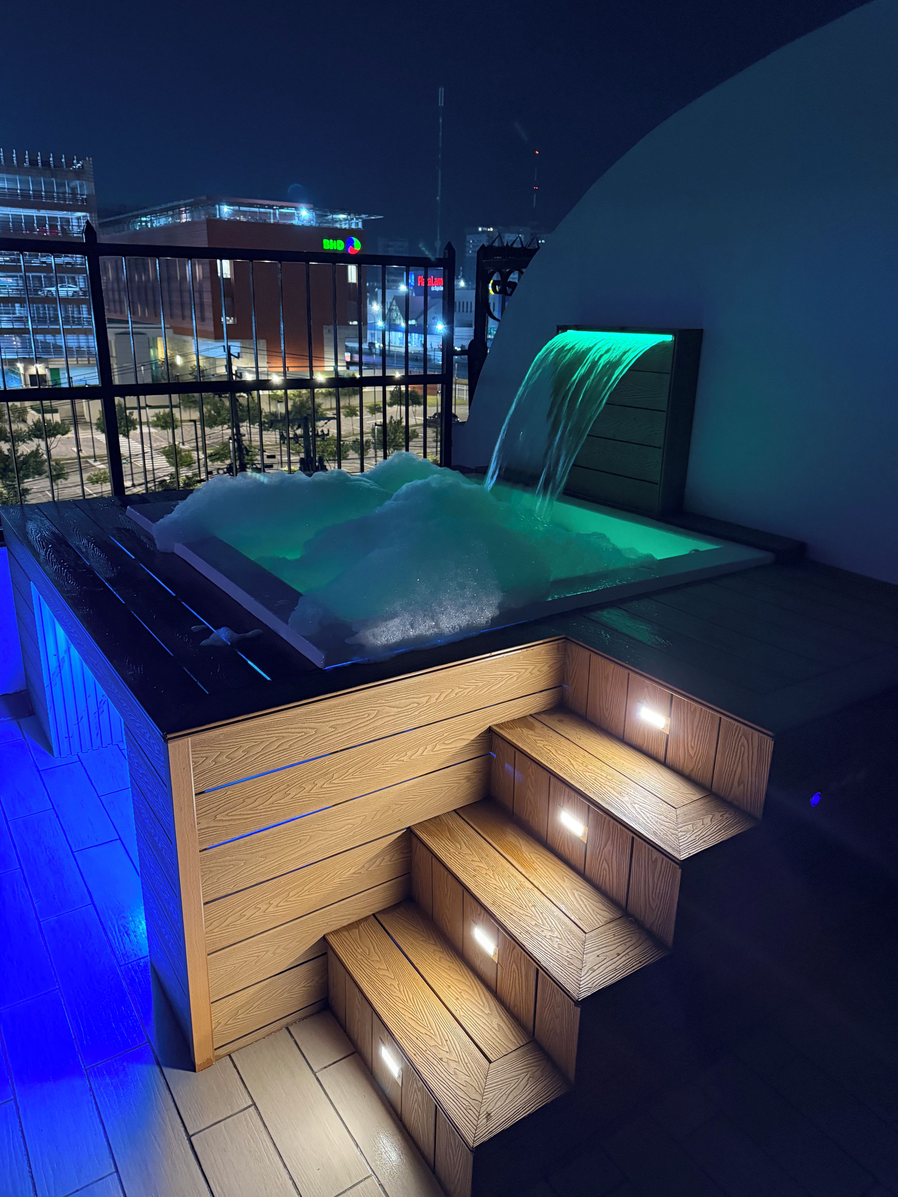 Un bain à remous doté d’une cascade, d’un escalier en bois et offrant une vue sur la ville la nuit.