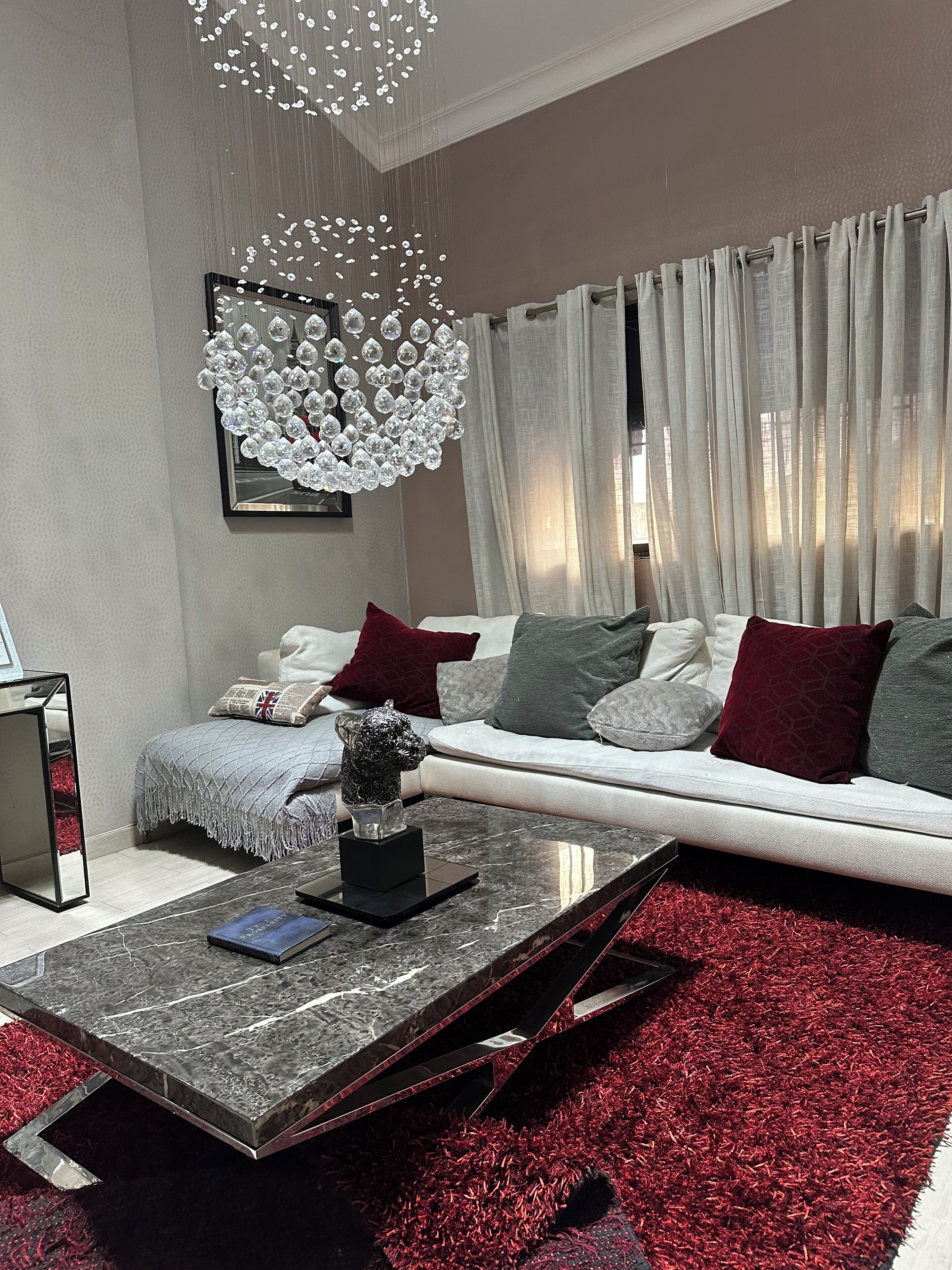 Ein modernes Wohnzimmer mit einer Couch, roten und grauen Kissen, einem Couchtisch mit einem Buch und einem dekorativen Gegenstand, einer Chandelüster und leichten Vorhängen.