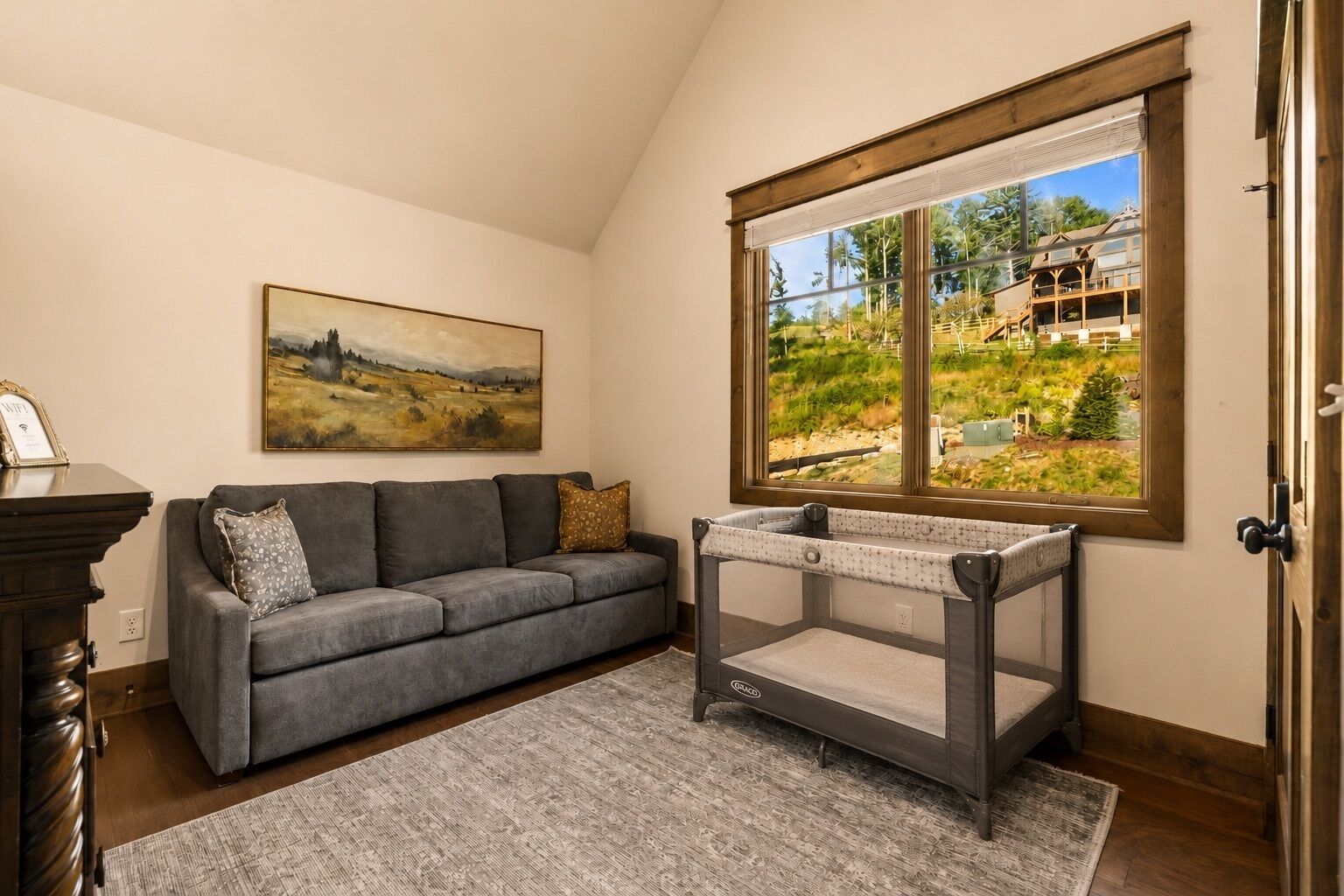 A room with a gray sofa, a painting, a crib, and a window with a view of a house and greenery.
