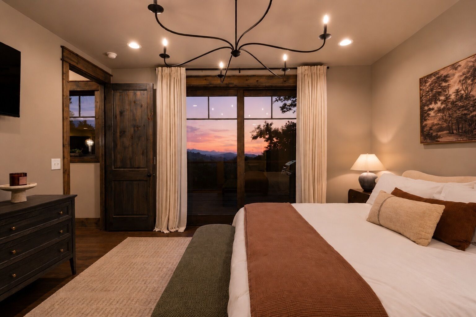 Een slaapkamer met een groot bed, een nachtkastje, een lamp, een dressoir en uitzicht op het buitenleven door een glazen deur.