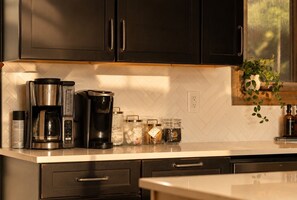 Start your morning at the coffee station, stocked with both a drip brewer and single-serve machine against a herringbone tile backsplash. Warm natural light pours in through the window above.