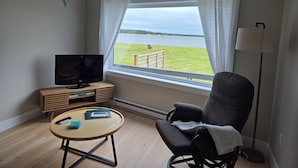 A room with a television, a small table with a book, a chair, and a view of a grassy area and water through a window.