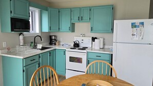 A compact kitchen with teal cabinets, a white refrigerator, a stove, and a microwave. There is a wooden dining table with chairs and a basket on it.