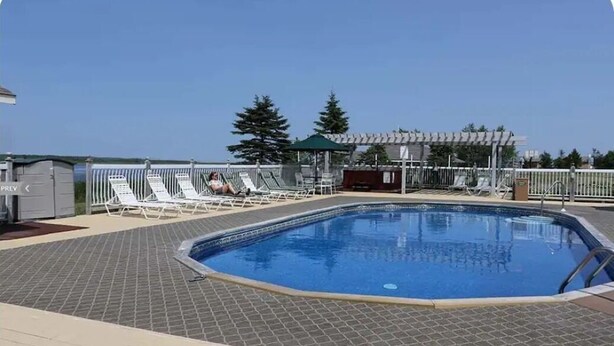 A rectangular swimming pool with a tiled deck, surrounded by white lounge chairs and a covered seating area.