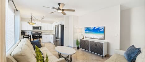 A modern living room with a sofa, a small round table, a TV mounted on the wall, and a kitchen area in the background.