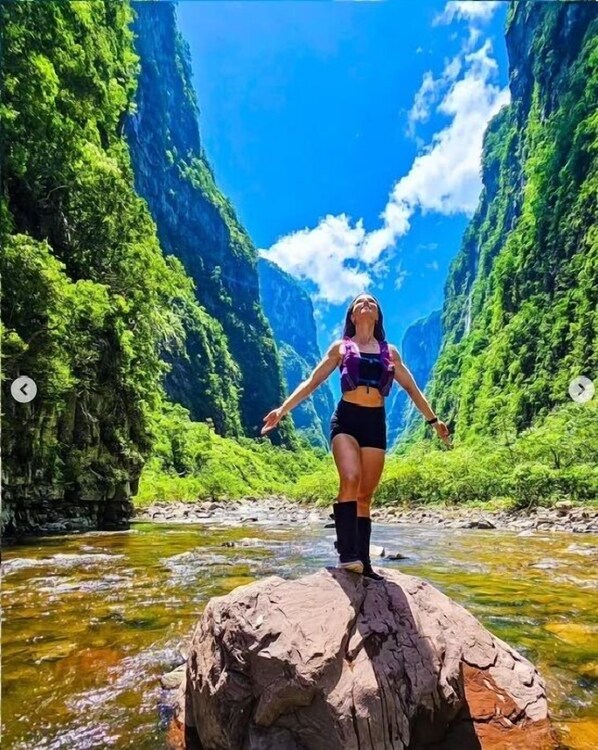 Une personne se tient sur un rocher au bord d’une rivière, avec une végétation luxuriante et des montagnes en arrière-plan.