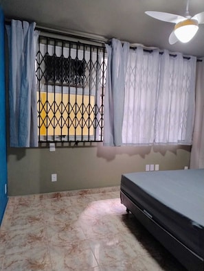 Un dormitorio con una cama, ventilador de techo, ventana con cortinas y suelo de baldosas.