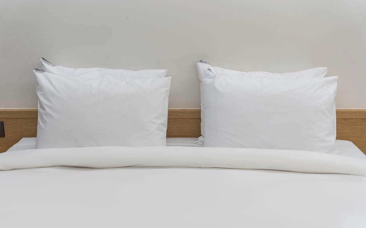 A bed with white bedding and pillows, a wooden headboard, and a plain wall.