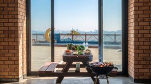 A patio with a grill, picnic table, and food items, overlooking a waterfront with a city skyline. - Red Sky Daebudo (Ansan)