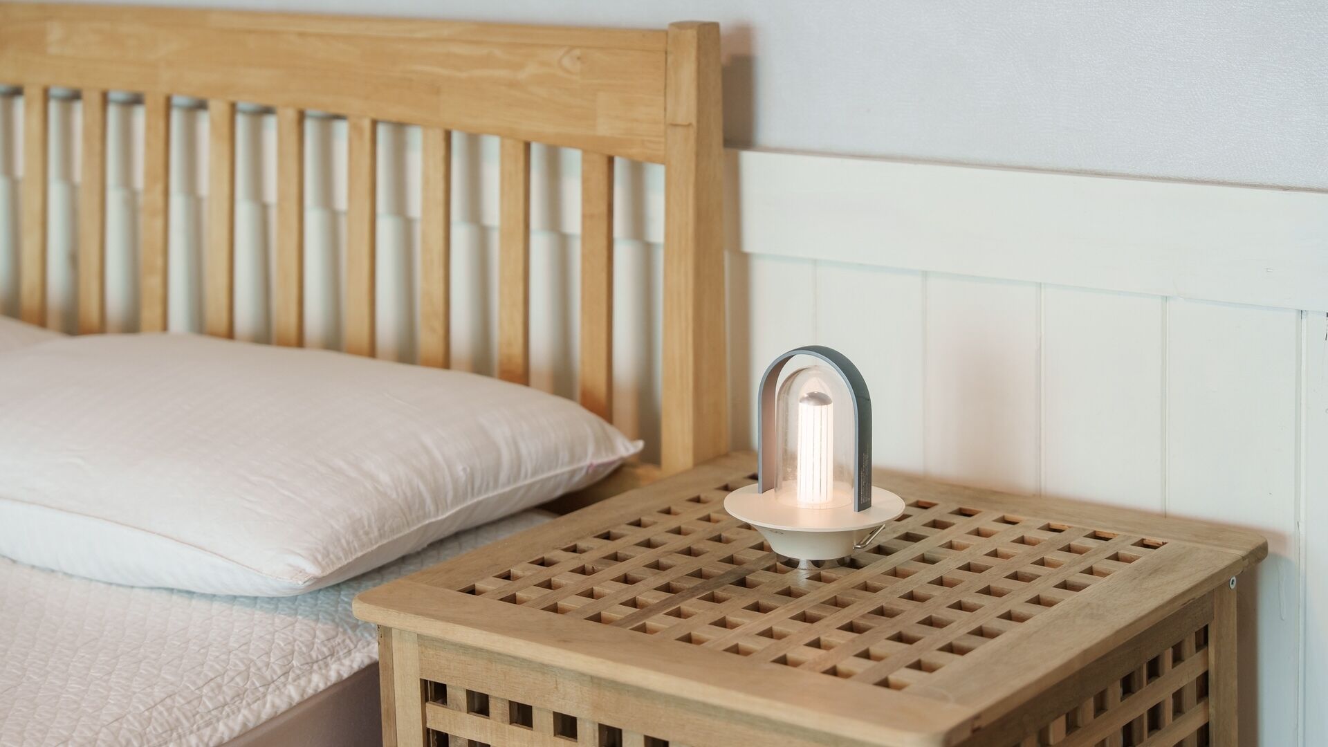 Ein Nachttisch aus Holz mit einer Lampe, ein Bett mit einem Holz-Kopfteil und eine weiße Wand.
