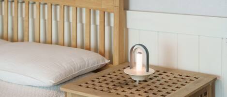 Ein Nachttisch aus Holz mit einer Lampe, ein Bett mit einem Holz-Kopfteil und eine weiße Wand.