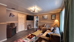A living room with a wooden floor, a sofa set, a television, and a fireplace. - Charming 5-bedroom house in Jackman / Moose River perfect for families, groups. (Moose River)