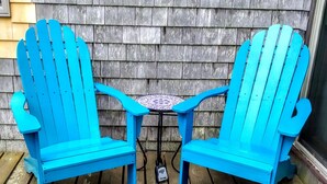 Deux chaises Adirondack bleues et une petite table ronde surmontée d’une mosaïque, disposées sur une terrasse en bois, adossées à un mur en bois.
