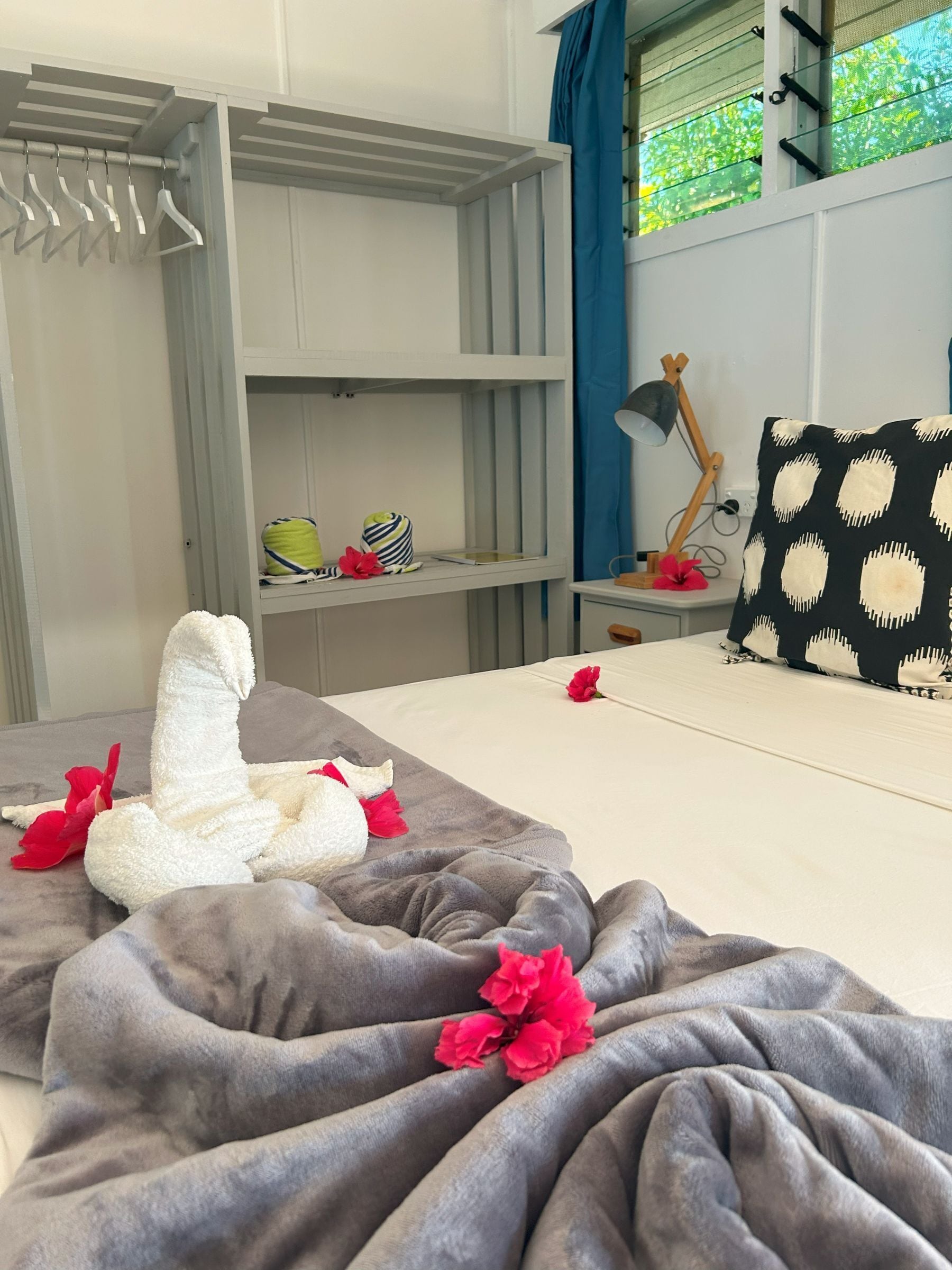 A neatly made bed with a grey blanket and red flowers, a grey wardrobe with hangers, a small shelf with a bag and a hat, and a window with blue curtains.