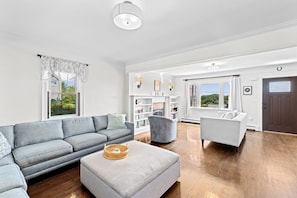 Ein geräumiges Wohnzimmer mit einem grauen Sofa, einem grauen Sessel und einem weißen Hocker. Ein Bücherregal mit Dekorationselementen und ein großes Fenster mit Vorhängen.