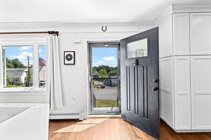 Ein Zimmer mit Holzboden, weißen Wänden, einer weißen Tür und Blick auf eine Straße und Häuser.