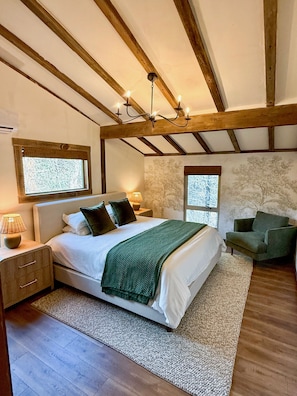 A bedroom with a large bed, wooden beams, a chandelier, and two windows.