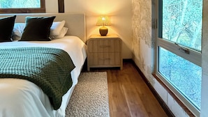 Ein Schlafzimmer mit Bett, Nachttisch mit Lampe, Fenster mit Aussicht und Holzbalken an der Decke.