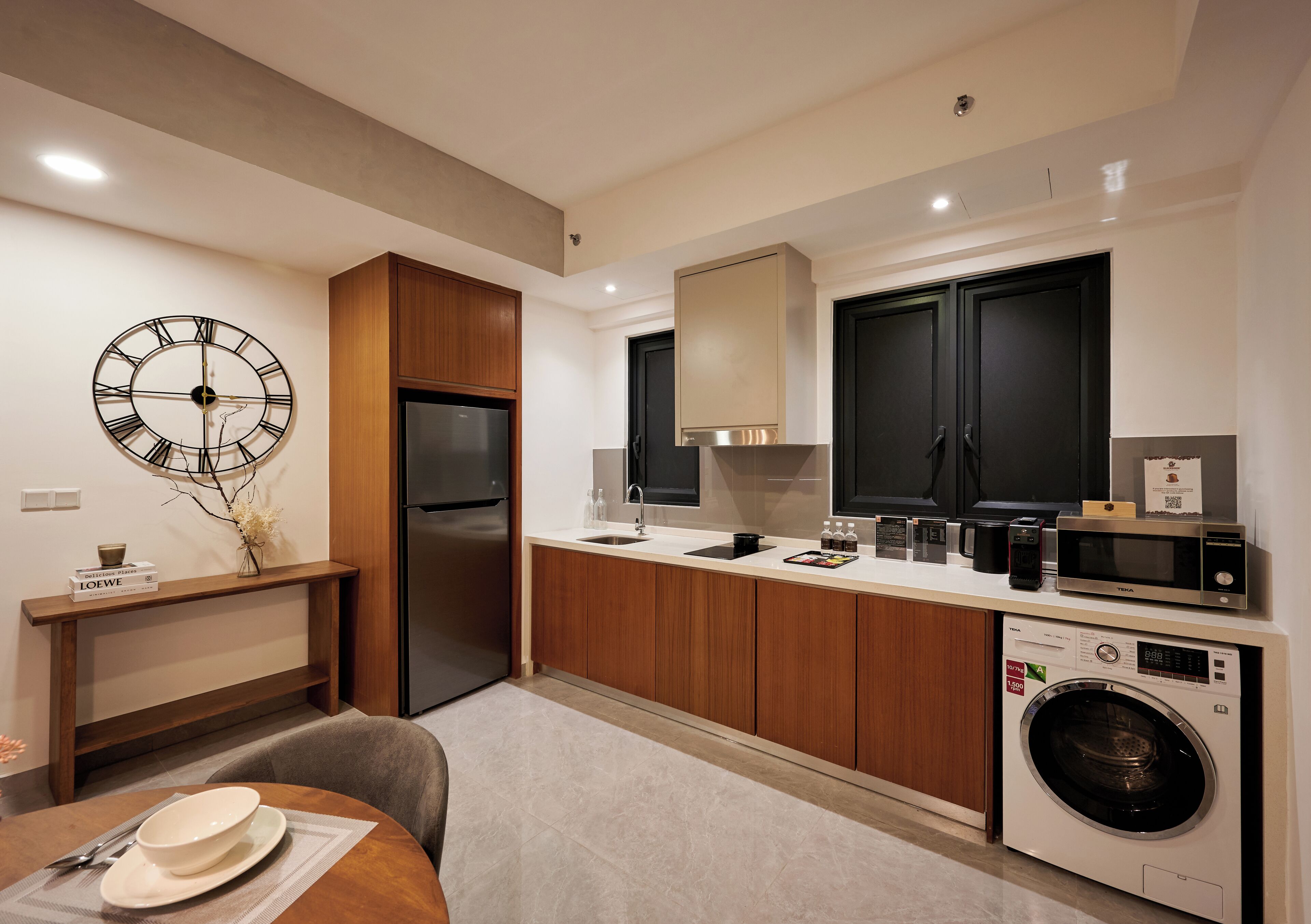 A modern kitchen with wooden cabinets, a built-in washing machine, and a large wall clock.
