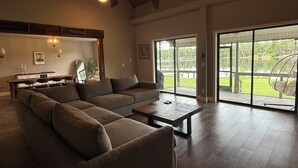 A spacious living room with a sectional sofa, a coffee table, and a dining area with a bench. Large sliding glass doors lead to a deck with a view of a lake and trees. - Lakefront Retreat Tampa, 7BR, Dock & Fun (Lutz)