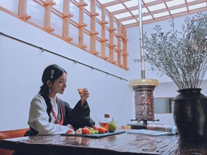 A person sitting at a table with a variety of fruits and a drink, in a room with a wooden ceiling structure and a potted plant.