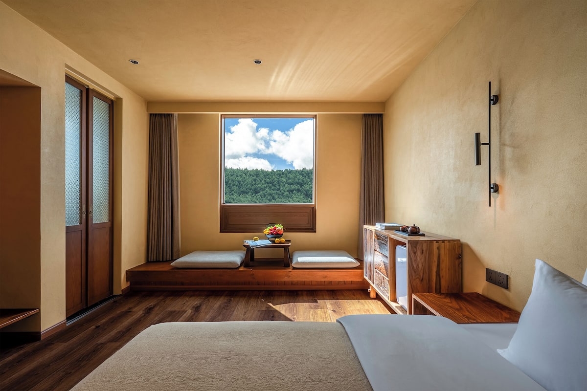 Quarto com uma cama grande, um banco de madeira, uma mesa pequena com frutas e uma janela com vista para uma floresta.