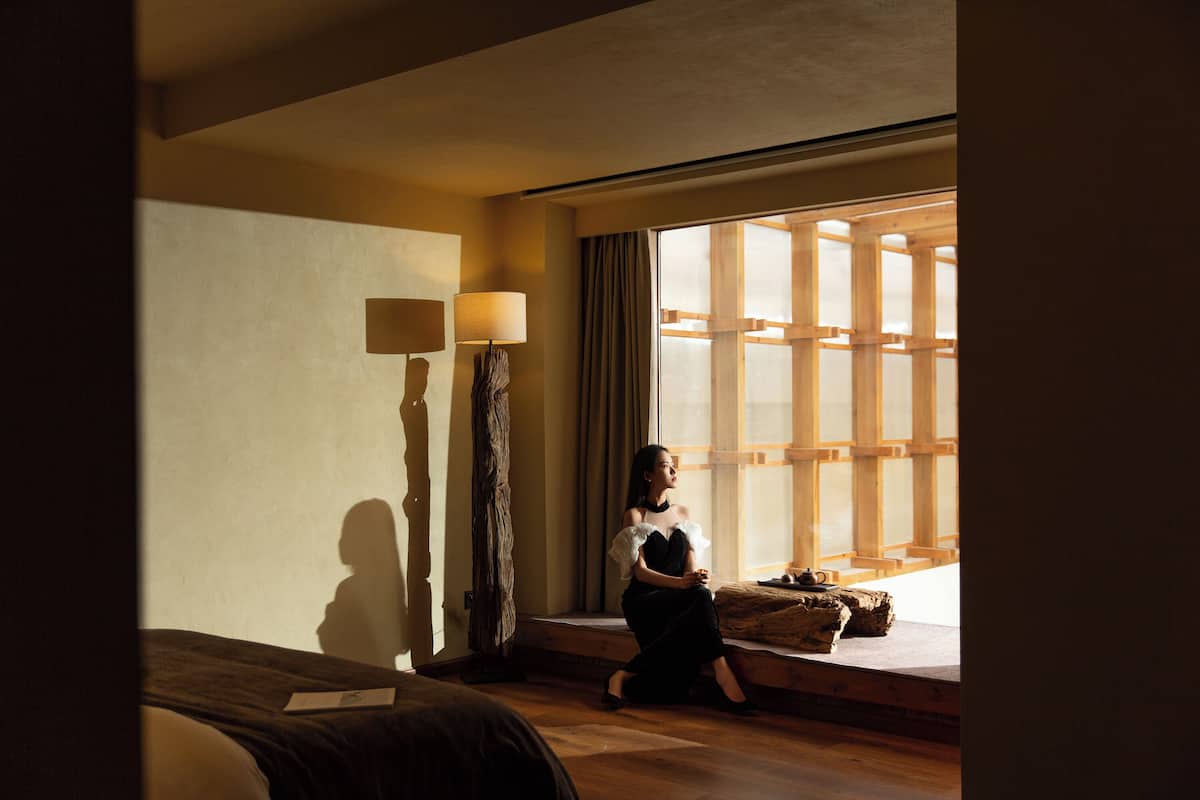 Quarto com cama, janela com persianas de madeira, luminária e uma pequena mesa com chaleira.
