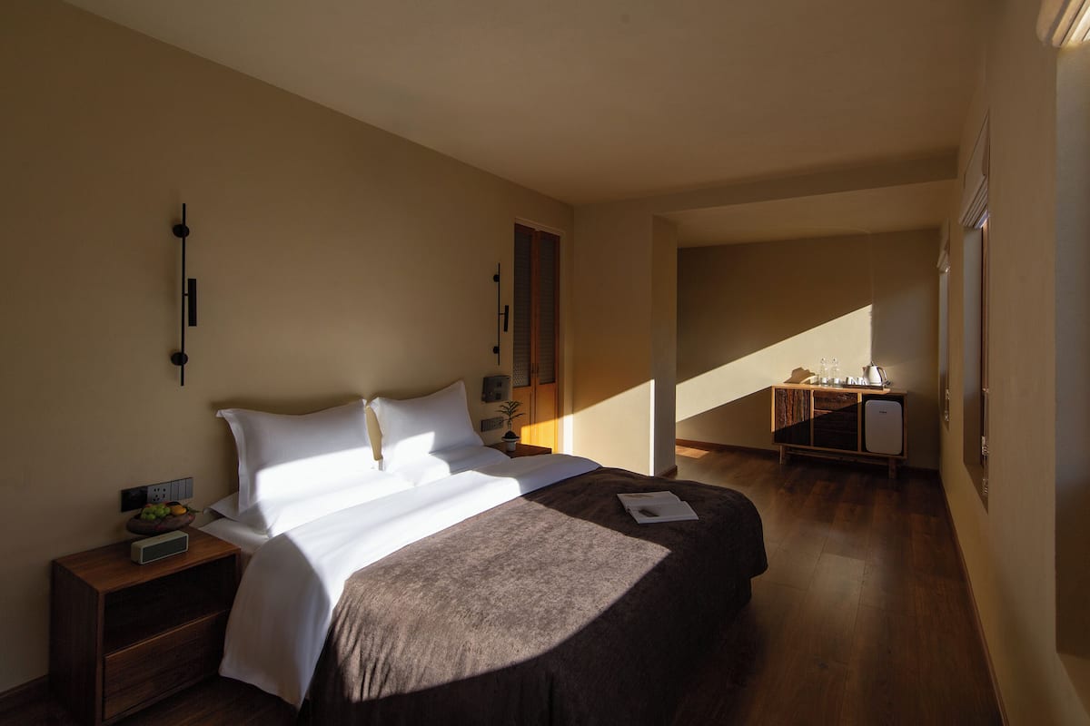 Quarto de hotel com uma cama grande, mesa de cabeceira com abajur, pequena mesa com uma planta e escrivaninha com computador.