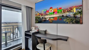 A modern hotel room with a large TV displaying a colorful amusement park, a balcony with a view of the park, a desk with a chair, and a small table with a coffee cup and bottles. - WEN SI TE HOTEL  (Shanghai)
