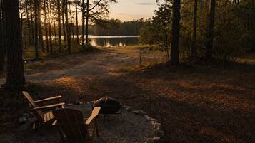 A campsite with two Adirondack chairs, a fire pit, and a lake in the background.