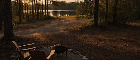 Een camping met twee Adirondack stoelen, een vuurkuil en een meer op de achtergrond.