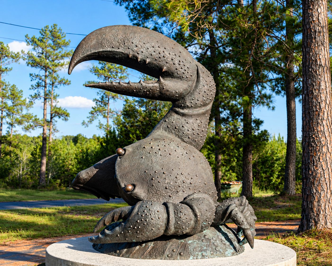 A sculpture of a large, textured creature with a prominent mouth and claws, set on a circular base in a natural setting with trees and a clear sky.