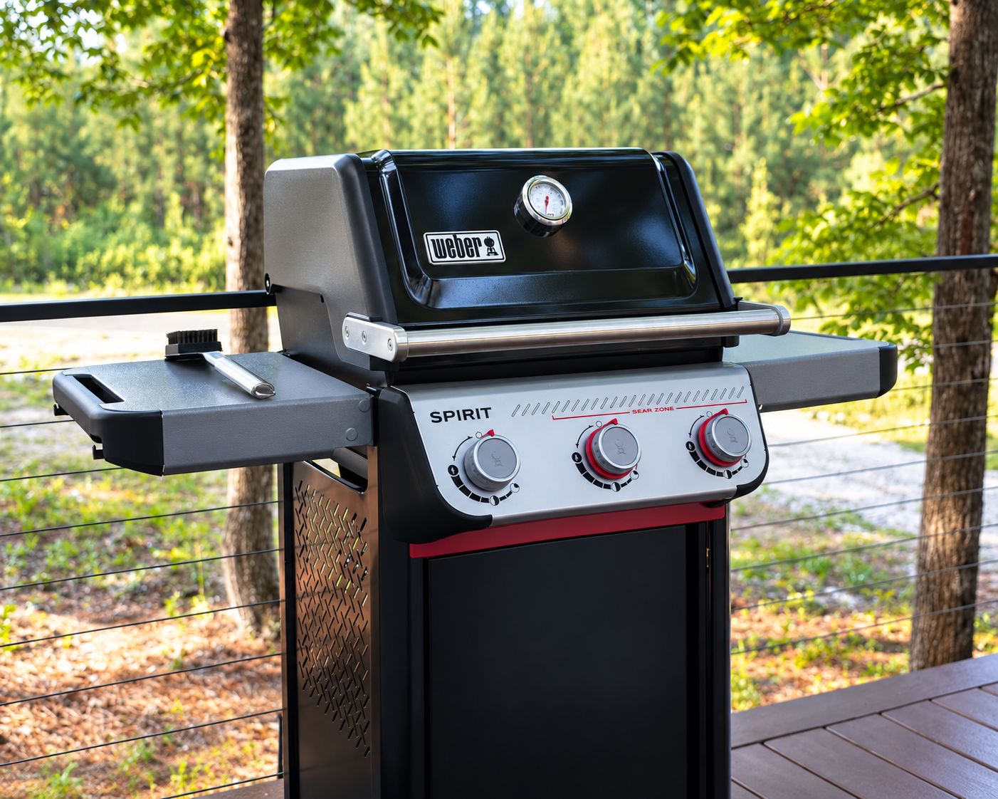 A Weber Spirit grill with a digital thermometer and a side table, set outdoors with a wooden deck and a forest background.