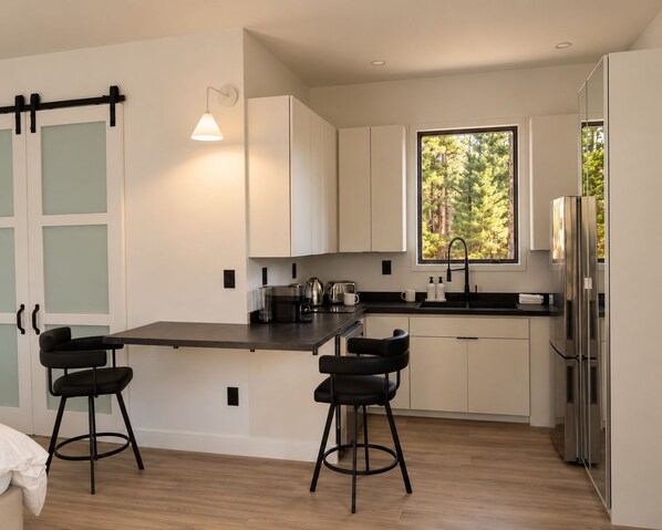 A modern kitchen with a central island, two bar stools, a window with a forest view, and a stainless steel refrigerator.