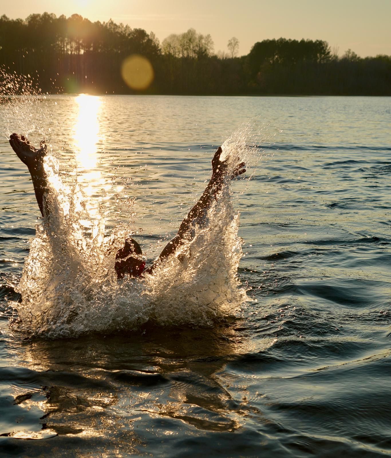 A person in the water with a splash, sun in the background, and trees on the horizon.