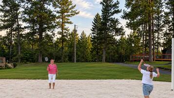 Two people playing badminton on a sandy surface with a grassy area and trees in the background.