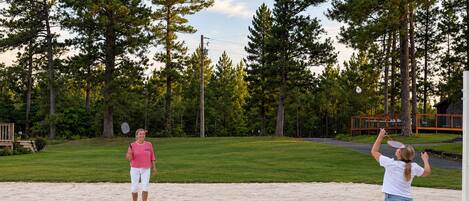 Two people playing badminton on a sandy surface with a grassy area and trees in the background.