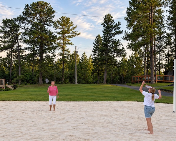 Two people playing badminton on a sandy surface with a grassy area and trees in the background.