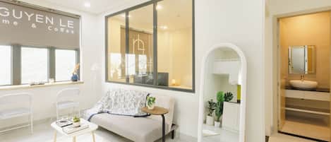 A modern hotel room with a sofa, a small table with a plant, a large mirror, and a window with a view of the outside.