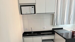 Μια μικρή, λειτουργική κου ζίνα με λευκά ντουλάπια, έναν μαύρο πάγκο και έναν εντοιχισμένο φούρνο μικροκυμάτων.